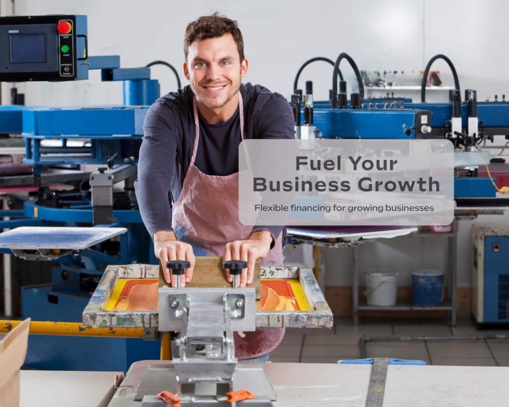 A small business owner operates a screen-printing machine in a workshop. Industrial equipment surrounds him as he smiles toward the camera in a manufacturing setting.