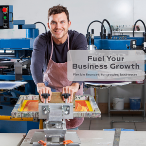 A small business owner operates a screen-printing machine in a workshop. Industrial equipment surrounds him as he smiles toward the camera in a manufacturing setting.